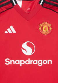 Rød Manchester United-drakt med v-hals, med svarte striper på skuldrene og hvit "Snapdragon"-logo i midten.