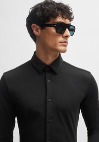 Schwarzes Langarmhemd mit Kragen und Knopfleiste. Der Stoff wirkt glatt und körperbetont. Schwarze quadratische Sonnenbrille ergänzt das Outfit.