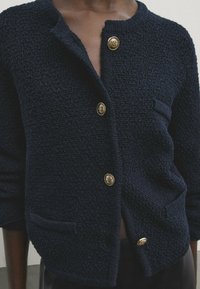 Marinblå texturerad cardigan med rund halsringning, prydd med fem gyllene knappar och två framfickor. Mjuk tyg, avslappnad passform och långa ärmar.