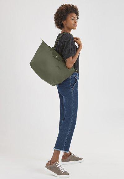 Olive green tote bag with rounded shape, button closure, and dual handles. Textured fabric with subtle logo detail. Worn on shoulder.