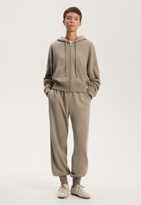 Felpa beige con cappuccio, zip e tasca frontale, abbinata a pantaloni sportivi a gamba affusolata e sneakers chiare.