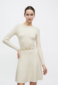 Πλεκτό εκρού φόρεμα με μακριά μανίκια, εφαρμοστή μπλούζα, φουσκωτή φούστα, υφασμένο διαμάντι μοτίβο και ταιριαστή ζώνη με χρυσή αγκράφα.