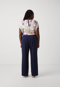 Le haut à fleurs Flo présente un motif abstrait coloré, des manches courtes et un dos en trou de serrure. Associé à un pantalon large bleu marine en tissu lisse.