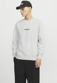 Sweat-shirt gris à col rond et manches longues, avec un petit logo noir sur la poitrine. Porté avec un pantalon noir et une casquette noire.