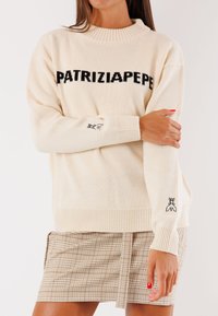 Maglione crema con testo nero "PATRIZIA PEPE", colletto e polsini a coste, con delicata ricamo di forbici sulla manica sinistra. Abbinato a una gonna scozzese.
