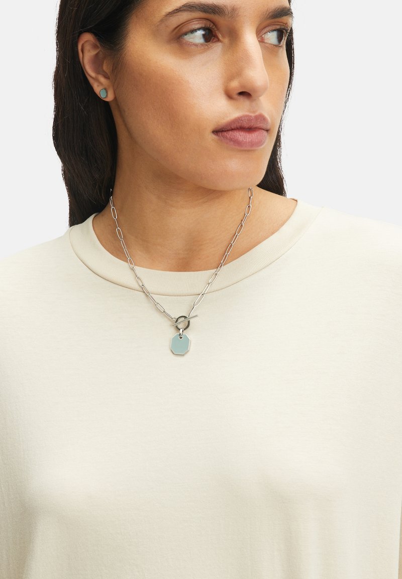 Zilveren ketting met een octagonale hanger en een ronde sluiting, gedragen over een lichtbeige, korthandsledige top. Minimalistisch ontwerp.