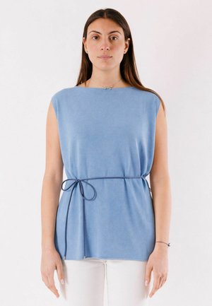 Donna con lunghi capelli castani che indossa un top azzurro senza maniche con una sottile cintura annodata e pantaloni bianchi, in piedi davanti a uno sfondo bianco.