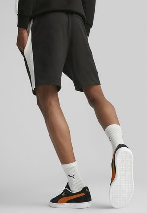 Sorte shorts med hvide sidelistrer, kombineret med sorte og orange sneakers og hvide ribbede sokker, hvilket fremhæver et afslappet atletisk design.