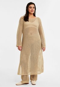 Beige Netzüberwurf mit langen Ärmeln, oversized Passform und offenem Strickmuster. Getragen über passenden beigen Hosen und beigen Schuhen.
