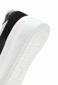 Zapatilla en blanco y negro con parte superior de ante negro texturizado, suela de goma blanca lisa y un patrón de agarre en la parte inferior.