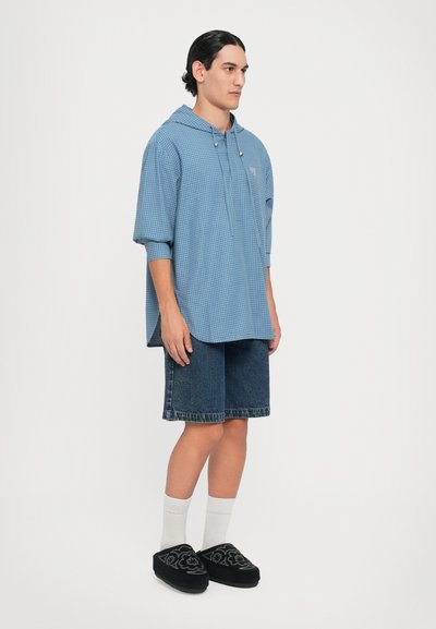 Camisa con capucha de gran tamaño a cuadros azules con un bolsillo frontal, combinada con pantalones cortos de mezclilla oscuros y zapatos negros sin cordones con bordados florales.