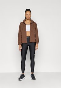 Καφέ ζακέτα με φερμουάρ και γιακά, φορεμένη πάνω από λευκό κοντό τοπ και μαύρο κολάν. Το σύνολο ολοκληρώνεται με μαύρα αθλητικά παπούτσια.