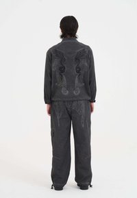 Grau Jacke mit Stickerei, die Flügel und Schlangen auf dem Rücken zeigt, kombiniert mit lockeren grauen Hosen; beide haben eine strukturierte Oberfläche.