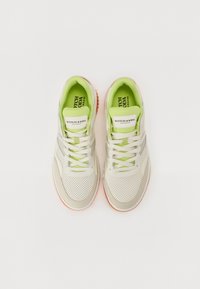 Sneaker mit einem Obermaterial in Beige und Grün, das eine perforierte Textur aufweist, weiße Schnürsenkel und eine gummierte orangefarbene Sohle hat. Markenlogo auf der Zunge sichtbar.