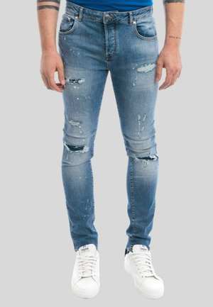 Jeans in denim blu sbiadito con strappi sfrangiati e macchie di vernice, caratterizzati da un design aderente e cinque tasche, indossati con scarpe da ginnastica bianche.
