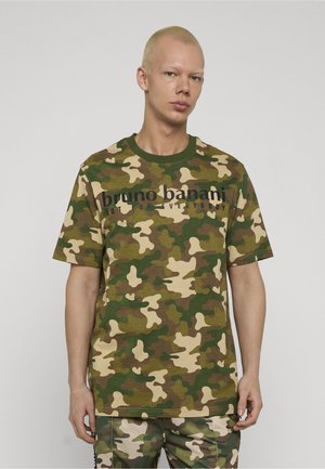 Persona con capelli biondi corti che indossa una maglietta e pantaloni mimetici verdi e marroni, in piedi contro uno sfondo chiaro e uniforme.