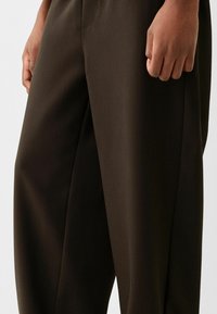 Bruna skräddarsydda byxor av ett slätt tyg. Har en avslappnad passform och lätt veck fram, med fickor på vardera sida.