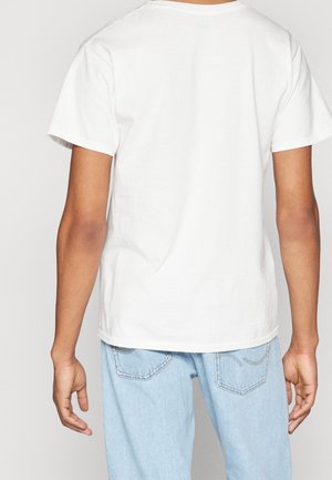 Persona che indossa una t-shirt bianca a maniche corte semplice e jeans azzurri chiari, vista da dietro su uno sfondo neutro.