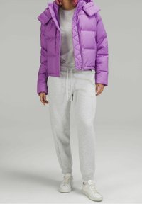 Veste gonflée violette avec capuche, design matelassé, associée à un pantalon de survêtement gris et des baskets blanches. La veste est munie de boutons et présente une coupe courte.