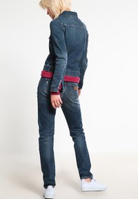 Syvän sininen farkkutakki, istuva malli etutaskuilla, yhdistetty tummansinisiiin farkkuihin ja valkoisiin lenkkareihin. Punainen paita näkyy sen alla.