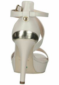 Sandal alto bianco con tacco, accentuato da dettagli metallici oro, dotato di una fascetta alla caviglia regolabile e un design elegante con piattaforma. Texture in pelle liscia.