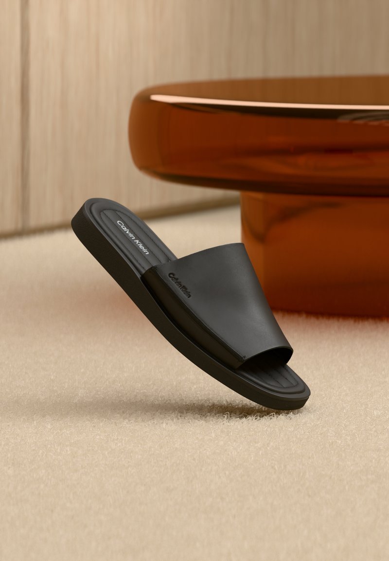 Sandale slide noire Calvin Klein reposant sur un tapis beige à côté d'une table ronde de couleur ambre, avec un mur en panneaux de bois en arrière-plan.