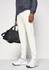 Vita atletiska byxor med ribbade muddar, i kombination med en mörkgrå jacka. Vita sneakers med blå accenter. Svart gymväska hålls i handen.