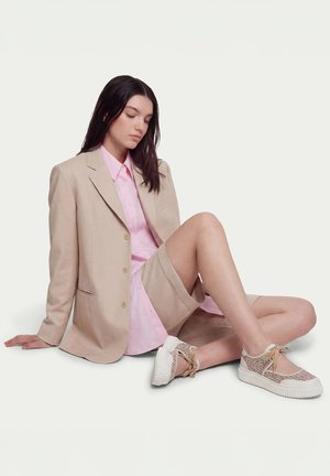 Young woman sits on floor wearing beige blazer, matching shorts, pink shirt, and beige patterned sneakers with thick white soles.