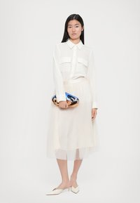 Transparante witte blouse met twee borstzakken, gelaagde tule rok, puntige witte flats en een glanzende blauw-gouden clutch.