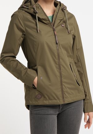 Femme portant une veste à capuche vert olive avec fermeture éclair à l'avant et main dans la poche, associée à un jean gris foncé.