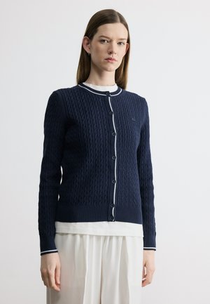 Žena stojaca pred bielym pozadím, oblečená v tmavomodrom pletenom kardigane so zapínaním na gombíky a manžetami s bielym lemom, ktorý má cez biely top a sukňu.