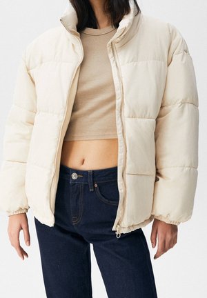Person trägt einen beigen Cropped-Top, eine cremefarbene, geöffnete Steppjacke und hoch taillierte dunkelblaue Jeans vor schlichtem Hintergrund.