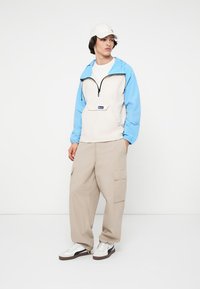 Junge Person in beigen Cargo-Hosen, weißen Sneakers, weißer Mütze und einer beigen sowie hellblauen Zip-Jacke, die vor einem weißen Hintergrund steht.