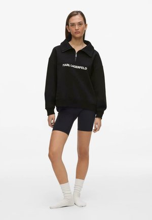 Kvinde iført sort Karl Lagerfeld sweatshirt med halv lynlås, sorte cykelshorts og hvide sokker, står med et neutralt udtryk.