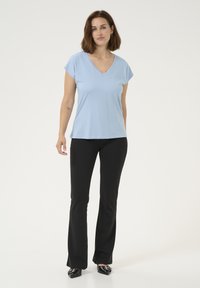 Un haut bleu clair en V avec des manches courtes, associé à un pantalon noir évasé et des chaussures noires, se tenant devant un fond blanc.