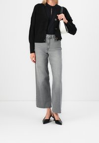 Schwarze gestrickte Cardigan, schwarze Bluse, hochtaillierte graue weite Jeans und schwarze spitze Absätze. Weiße Handtasche mit schwarzen Akzenten.
