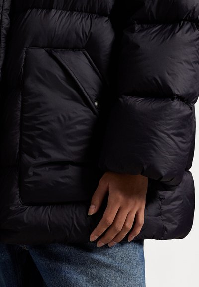 Marine blauwe pufferjack met een geplooide textuur, voorzien van een grote patchzak, een slanke pasvorm en een zachte glanzende afwerking. Hand rustend op het kledingstuk.