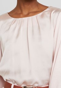 Helles rosa Satin-Oberteil mit weitem Ausschnitt, versehen mit Falten und luftigen Ärmeln. Es ist an der Taille mit einem braunen Gürtel und einer goldenen Schnalle gerafft.