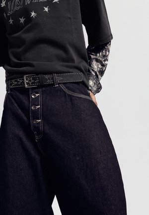 Persoană purtând blugi largi închiși la culoare cu nasturi în partea din față, o curea neagră cu ținte, un tricou negru cu stele și un tricou cu mânecă lungă cu model dedesubt.