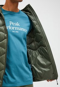 Quilted olive green jacket partially unzipped, revealing a teal t-shirt with white text "Peak Performance" and "Est. 1986." Smooth texture.