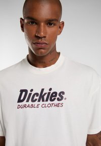 Bílé bavlněné tričko s výrazným tmavě červeným logem, na kterém je text "Dickies DURABLE CLOTHES" v jednoduchém písmu bez patkových písem.