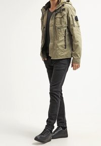 Olive green jacket with a hood, multiple pockets, and black accents. Worn over a dark top and paired with black slim-fit pants and sneakers.
