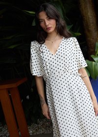 Femme aux longs cheveux foncés portant une robe blanche à pois noirs, debout près de plantes et de meubles en bois.
