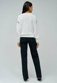 Sweat-shirt blanc à manches longues, col rond, avec des embellissements en bijoux aux épaules, associé à un jean taille haute foncé.