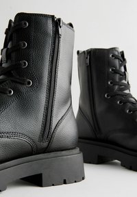 Zwarte leren legerlaarzen met een textuurafwerking, vetersluiting aan de voorkant, zijritsen en dikke rubberen zolen. Heeft metalen oogjes en gestikte accenten.