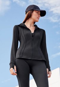 Crna atletska jakna s patentnim zatvaračem sprijeda i kapuljačom, s istaknutim šavovima. Uparen s crnim tajicama, nasuprot plavom nebu.