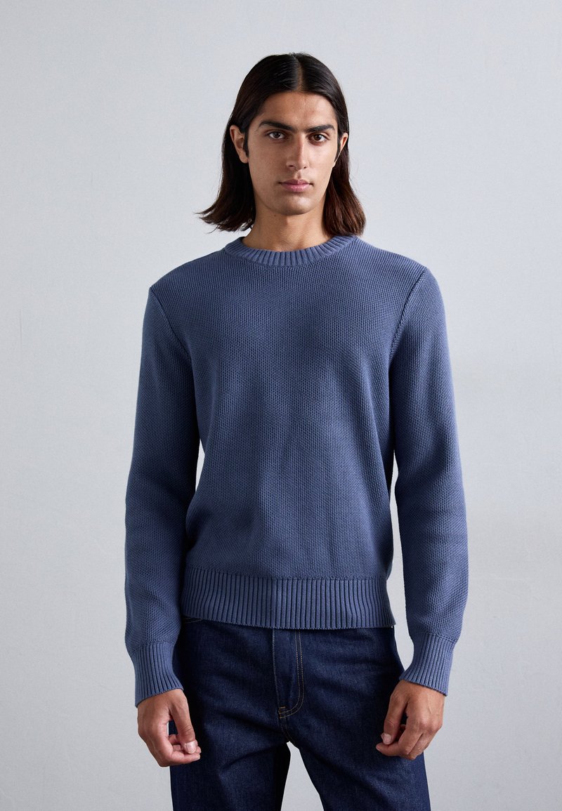 Blå strikket sweater med rund hals, struktureret mønster samt ribbede manchetter og kant, parret med mørke denimjeans. En simpel og moderne design.