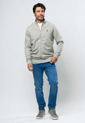 Férfi világosszürke cipzáras dzsekiben, fehér ingben, kék farmerral és szürke sportcipővel, egyszerű világos háttér előtt állva.