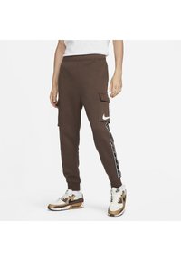 Bruna cargobyxor i sweatpants-modell med sidostriper, elastisk midja och flera fickor. Tillverkade av mjukt tyg, prydda med en vit Nike-logga.