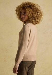 Bežs adīts džemperis ar garām piedurknēm, ribotām malām un pogu akcentiem uz piedurknēm, apvienots ar rakstainām biksēm un teksturētu fona. 